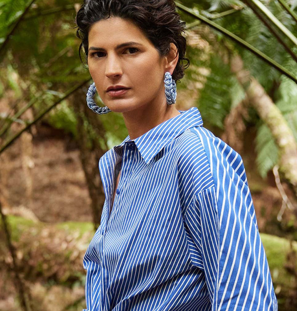 Close-up of a woman with short curly hair, wearing a striped blue shirt and large blue hoop earrings in a green natural setting