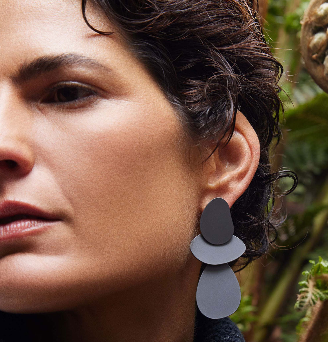 Close-up of a woman wearing large black geometric earrings, with soft curls framing her face, surrounded by greenery