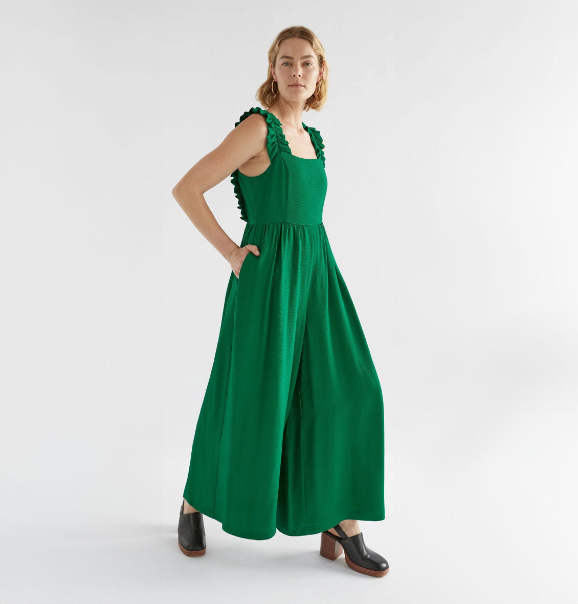 Model wearing a green sleeveless jumpsuit with ruffled straps and wide-leg design, styled with black heeled mules against a white backdrop